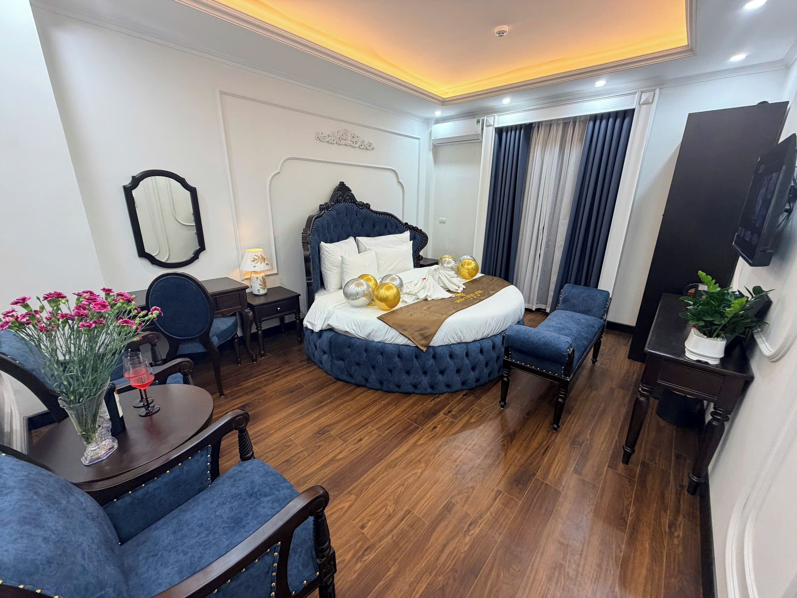 Habitación de hotel con una cama, dos sillones azules, un escritorio con lámpara, un espejo y una planta en maceta.