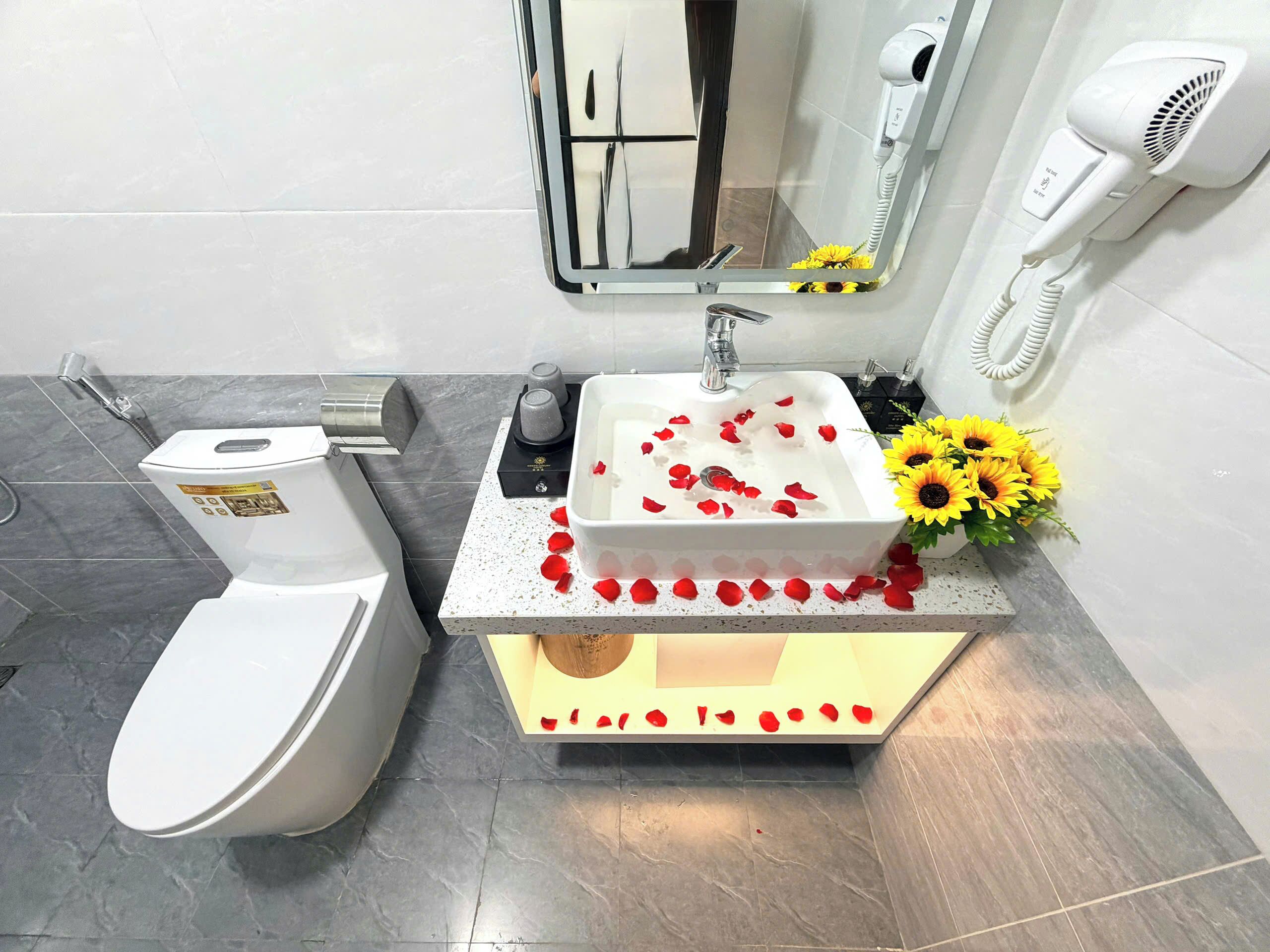 A bathroom with a white toilet, a sink with rose petals, a hairdryer, and a vase with sunflowers.