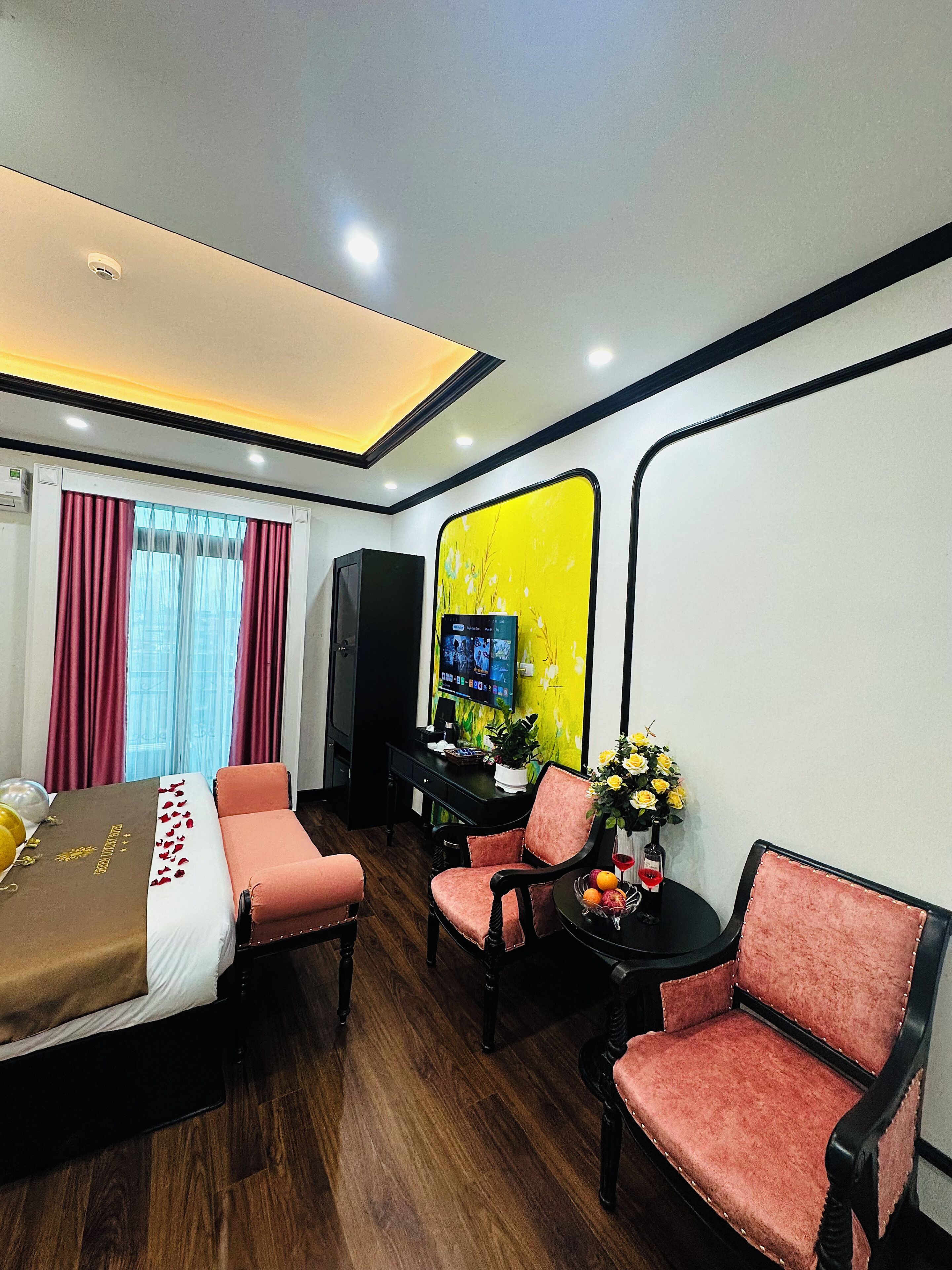 A hotel room with a bed, two pink chairs, a small table with a vase of flowers, a TV on a stand, and a yellow accent wall.
