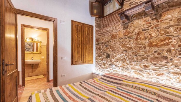 A bedroom with a stone wall, a wooden door, a striped bedspread, and a bathroom with a mirror and sink. - Domm'e Antoni - Apartment in the heart of Ogliastra in stone and juniper (Baunei)