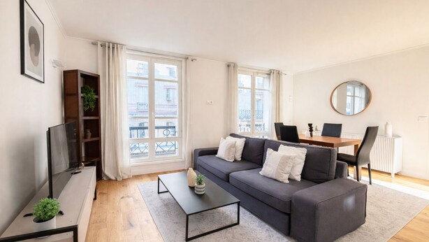 A modern living room with a grey sofa, a coffee table, a TV, and a dining area with chairs and a table. - Élégant 1 Chambre à Montparnasse (Paris)