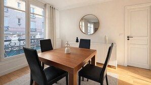 A dining room with a wooden table, black chairs, a round mirror, and a view of a balcony with a railing. - Élégant 1 Chambre à Montparnasse (Paris)