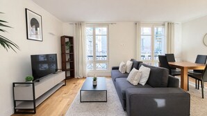 A modern living room with a flat-screen TV, grey sofa, wooden dining table, and large windows. - Élégant 1 Chambre à Montparnasse (Paris)