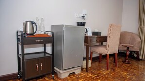 Een compacte hotelkamer met een kitchenette, een houten vloer, een bureau met stoel, en een klein kastje met waterkoker en glazen.