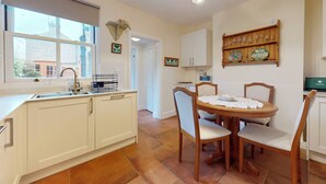 Una cocina con armarios blancos, una mesa de comedor con sillas, una ventana con vista a edificios y una repisa con platos y vasos.