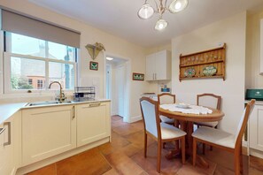 A kitchen with white cabinets, a dining table with chairs, a window with a view of buildings, and a shelf with plates and glasses. - Chic 2BR w/ Private Backyard - Near Loughton Tube (Loughton)