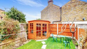 Una casita de juegos de madera con puertas de cristal, un jardín pequeño con un sendero empedrado y una valla de madera.