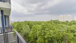 Un immeuble d’habitation doté d’un balcon donnant sur un feuillage verdoyant et luxuriant, ainsi que sur des gratte-ciel lointains, le tout sous un ciel nuageux.