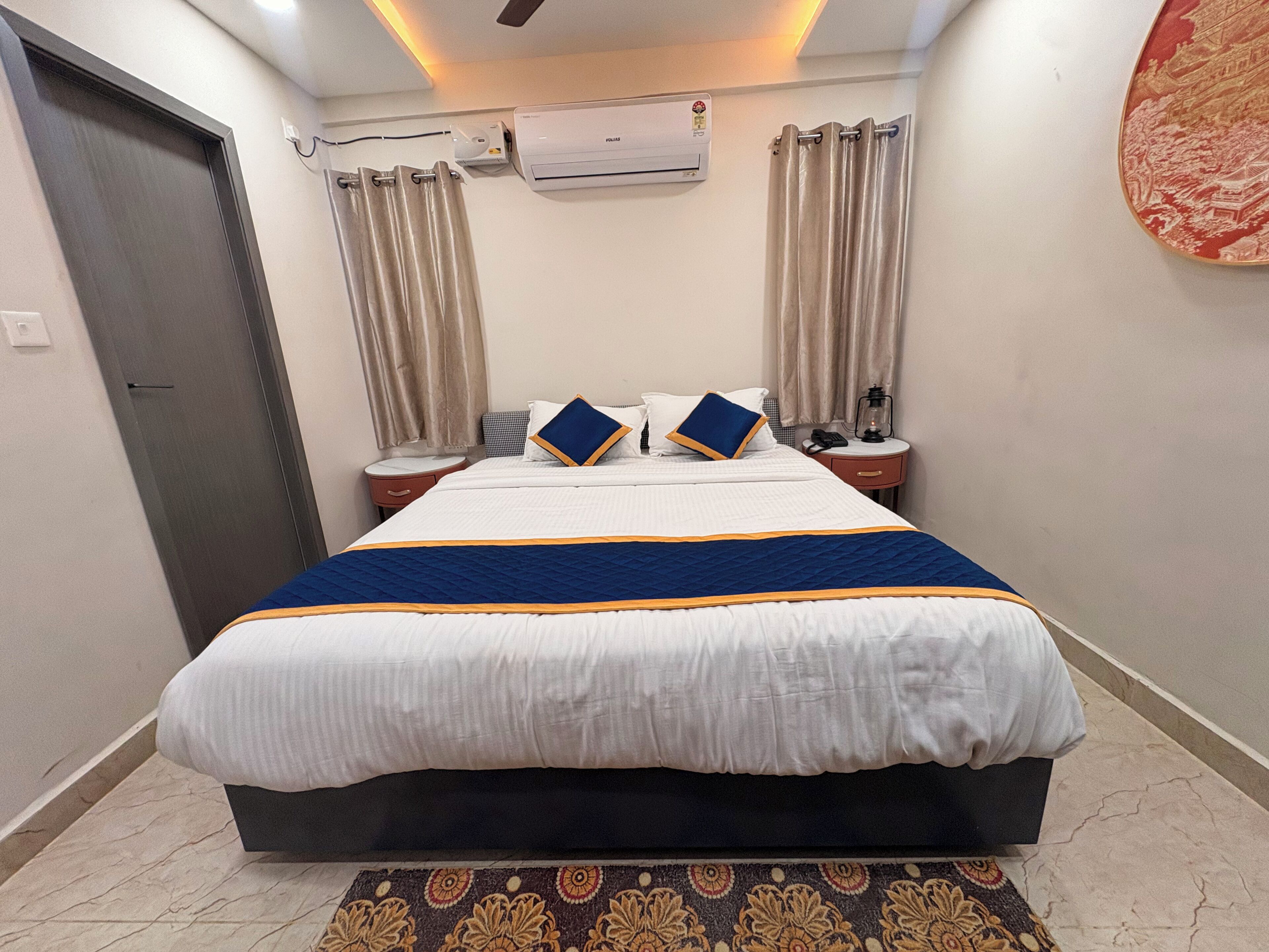 Una cama bien hecha con colcha azul y blanca, dos mesitas de noche con lámparas, un aire acondicionado de pared y una alfombra estampada.