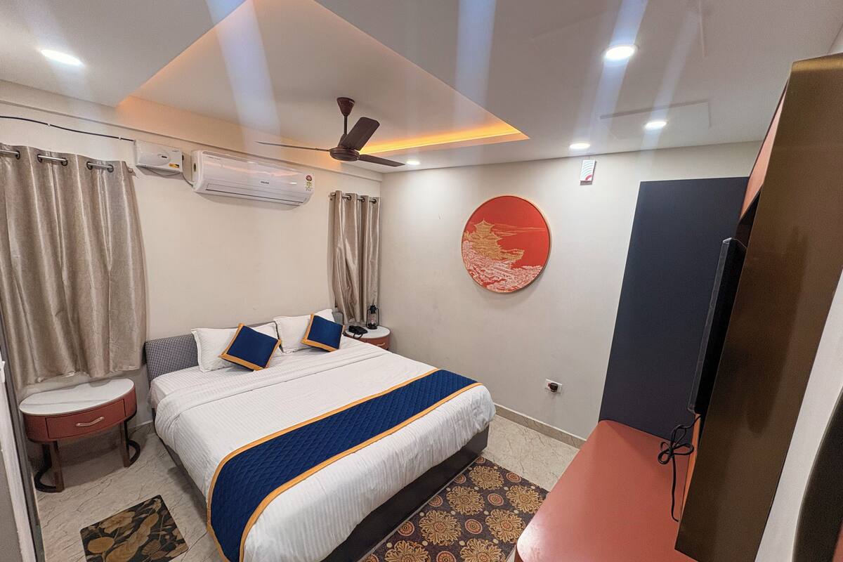 Habitación de hotel con cama, mesita de noche, aire acondicionado, cortinas, televisor montado en la pared y alfombra estampada.