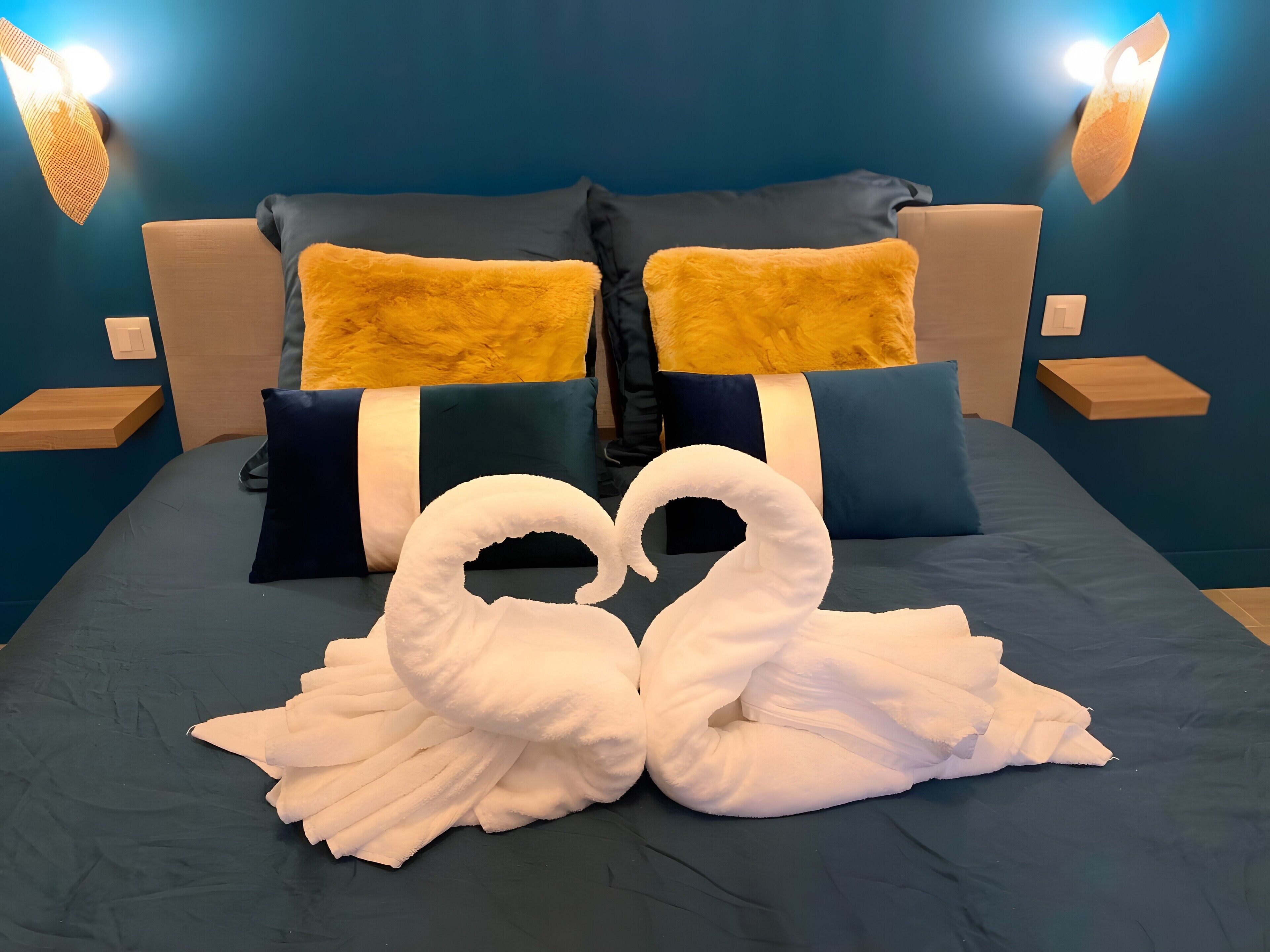 A bed with a blue bedspread, two pillows, and a towel arrangement in the shape of swans.