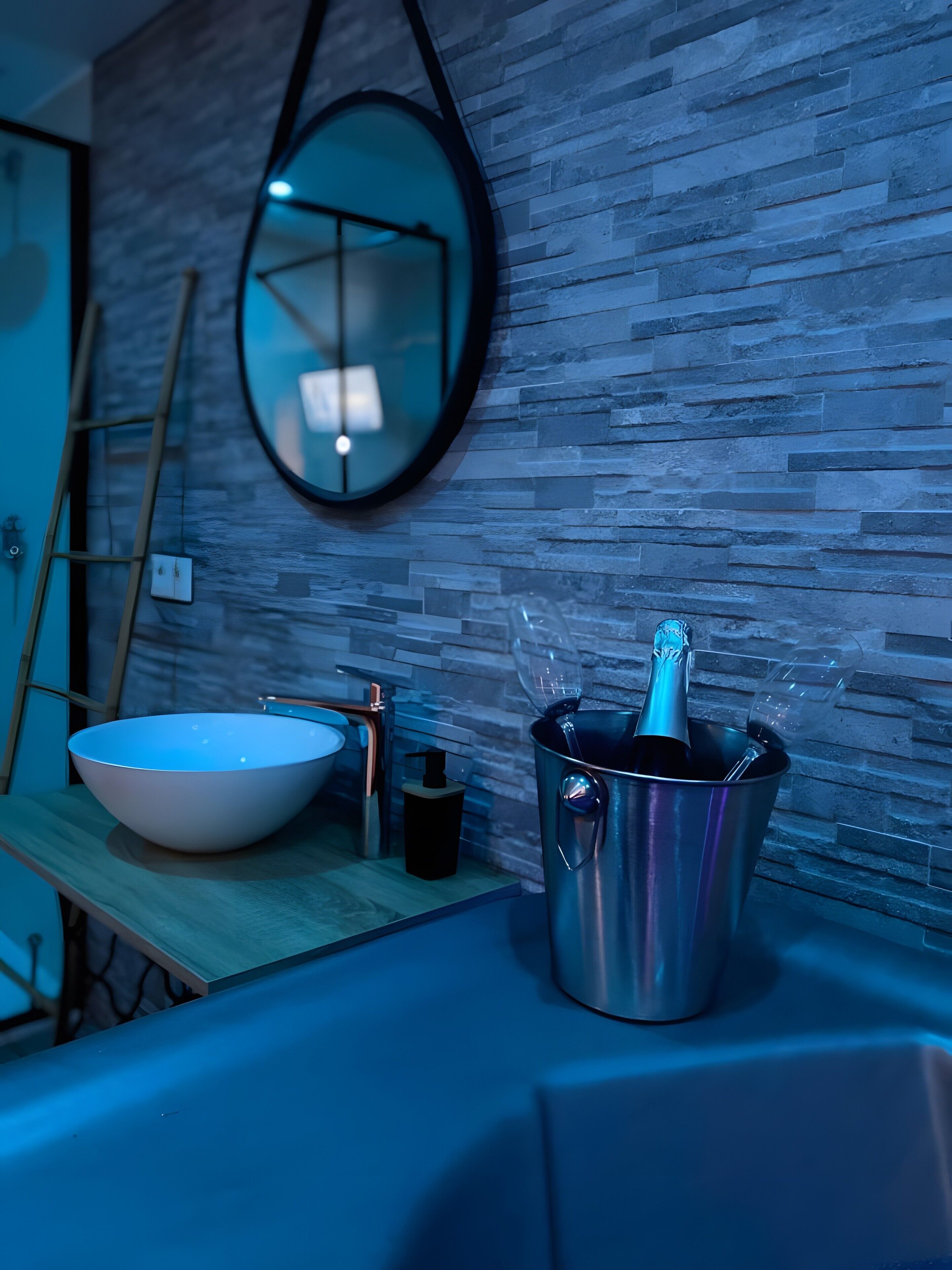 Une salle de bain avec un lavabo blanc, un miroir et un seau métallique contenant une bouteille.