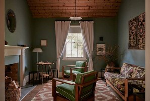 Un salon chaleureux avec une cheminée, un fauteuil vert, un canapé à motifs floraux et une fenêtre avec des rideaux.
