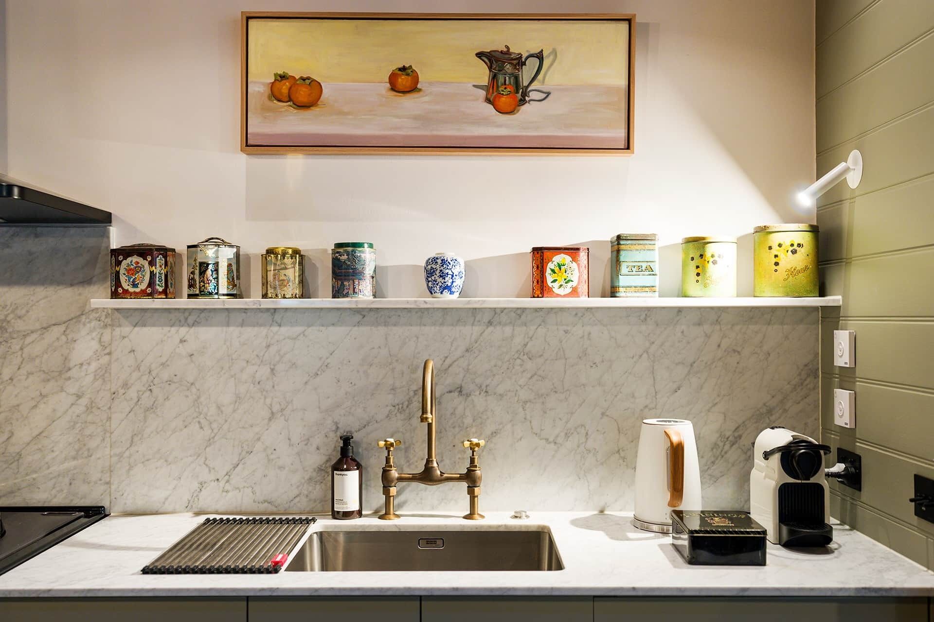 Una cocina con encimera de mármol, un lavamanos con grifo dorado, una cafetera y una estantería con varios recipientes.