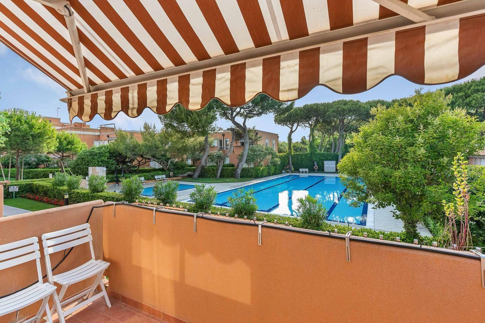A balcony with a striped awning overlooking a swimming pool, surrounded by greenery and buildings.