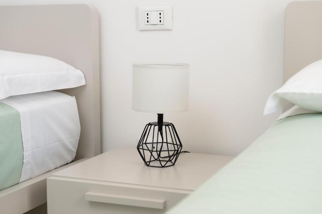 Una cama individual bien hecha con una colcha blanca y verde, una mesita de noche con una lámpara de estructura metálica negra y un cajón blanco.