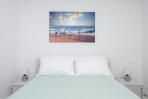 Une chambre à coucher avec un lit, des oreillers blancs et un tableau représentant une scène de plage, accroché au mur.