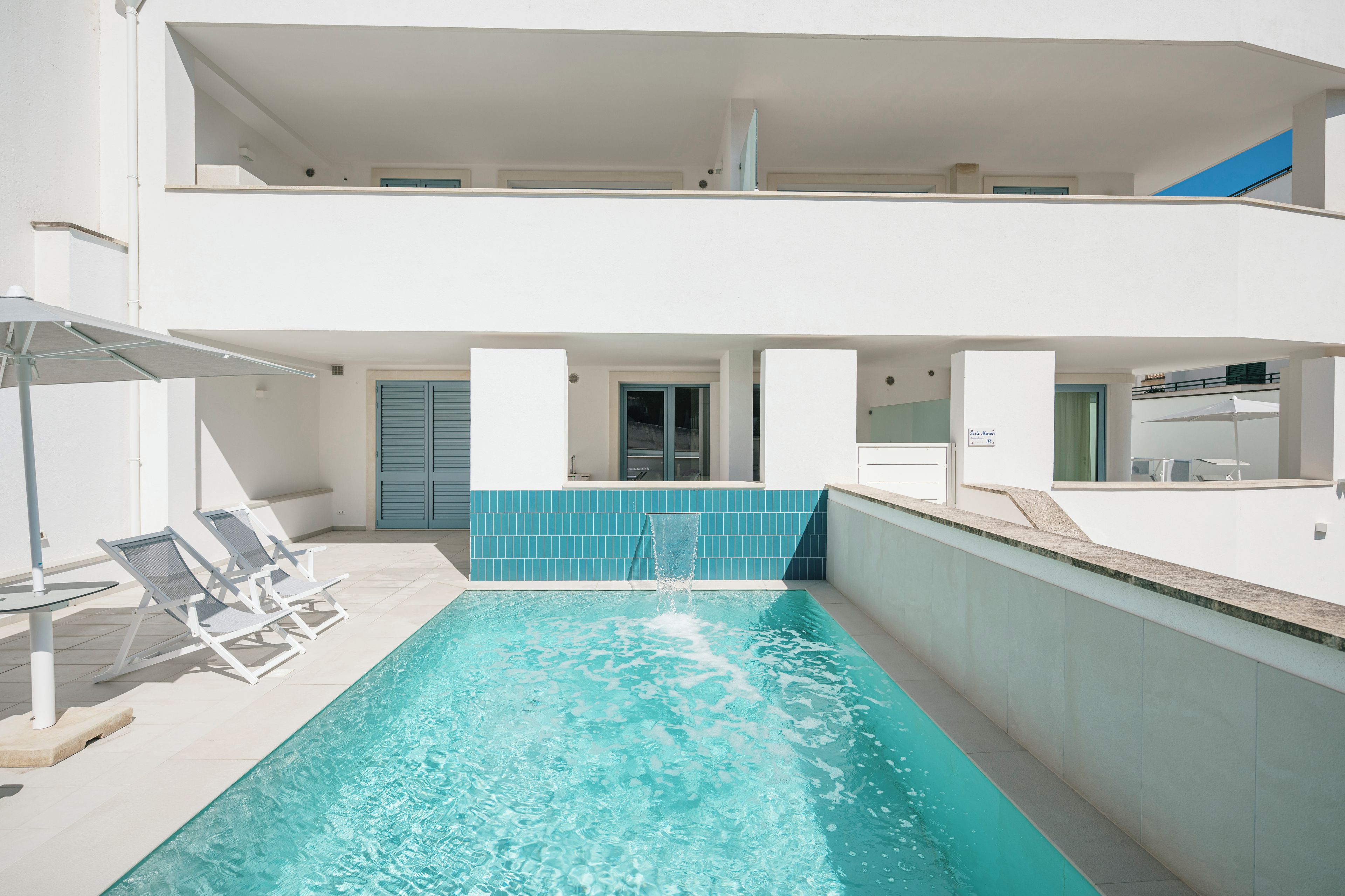 A swimming pool with a blue tiled wall, lounge chairs, and a sun umbrella.