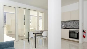 Cozinha moderna com armários brancos e painel de azulejos, área de refeições com mesa e cadeiras, e uma porta de vidro deslizante para uma área externa.