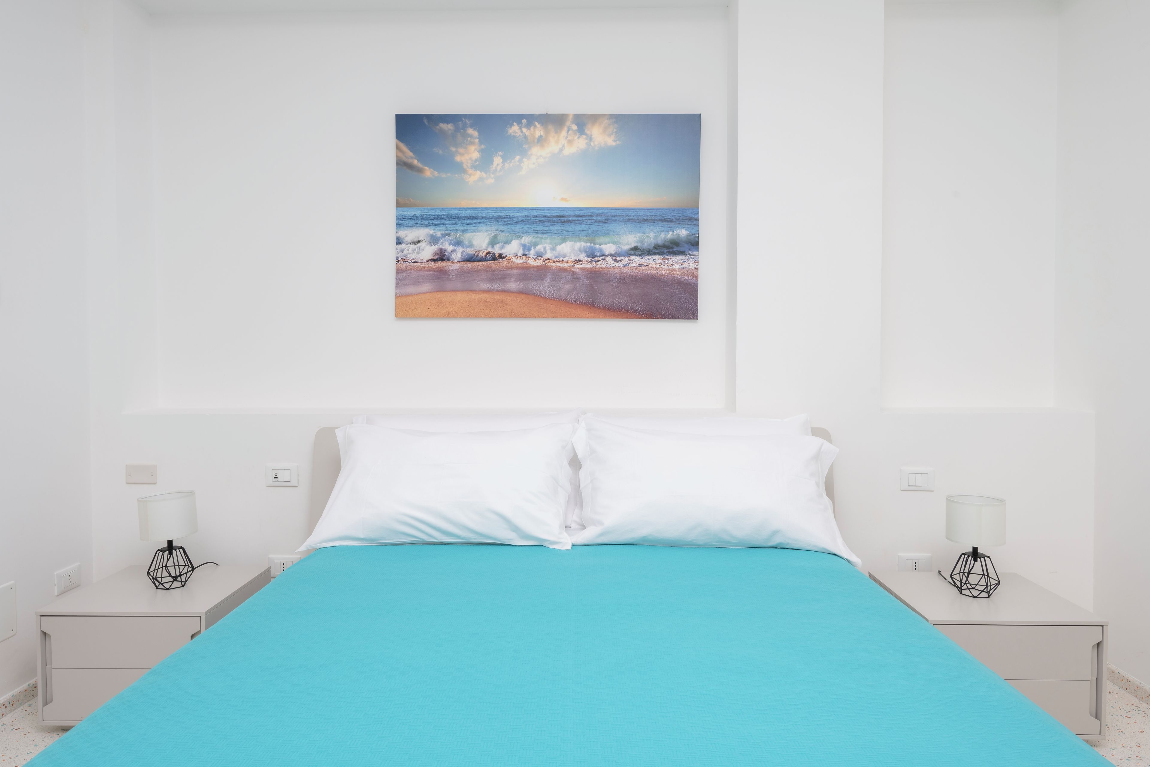 Une chambre à coucher avec un lit, des murs blancs, un tableau représentant une plage et deux tables de chevet avec des lampes.