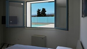 Quarto de hotel com cama, janela vista para piscina e árvores, e pequeno aquecedor no chão.