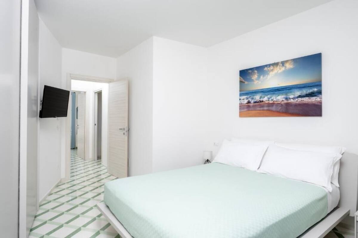 A bedroom with a bed, a painting of a beach scene, and a television on the wall.