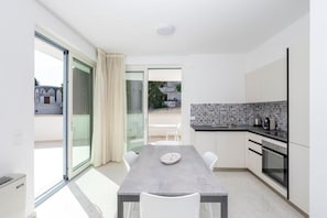 Une cuisine moderne avec un plan de travail gris, des armoires blanches et un pare-feu en carrelage. On y trouve une table à manger avec des chaises, ainsi qu’une porte-fenêtre coulissante donnant sur un espace extérieur.