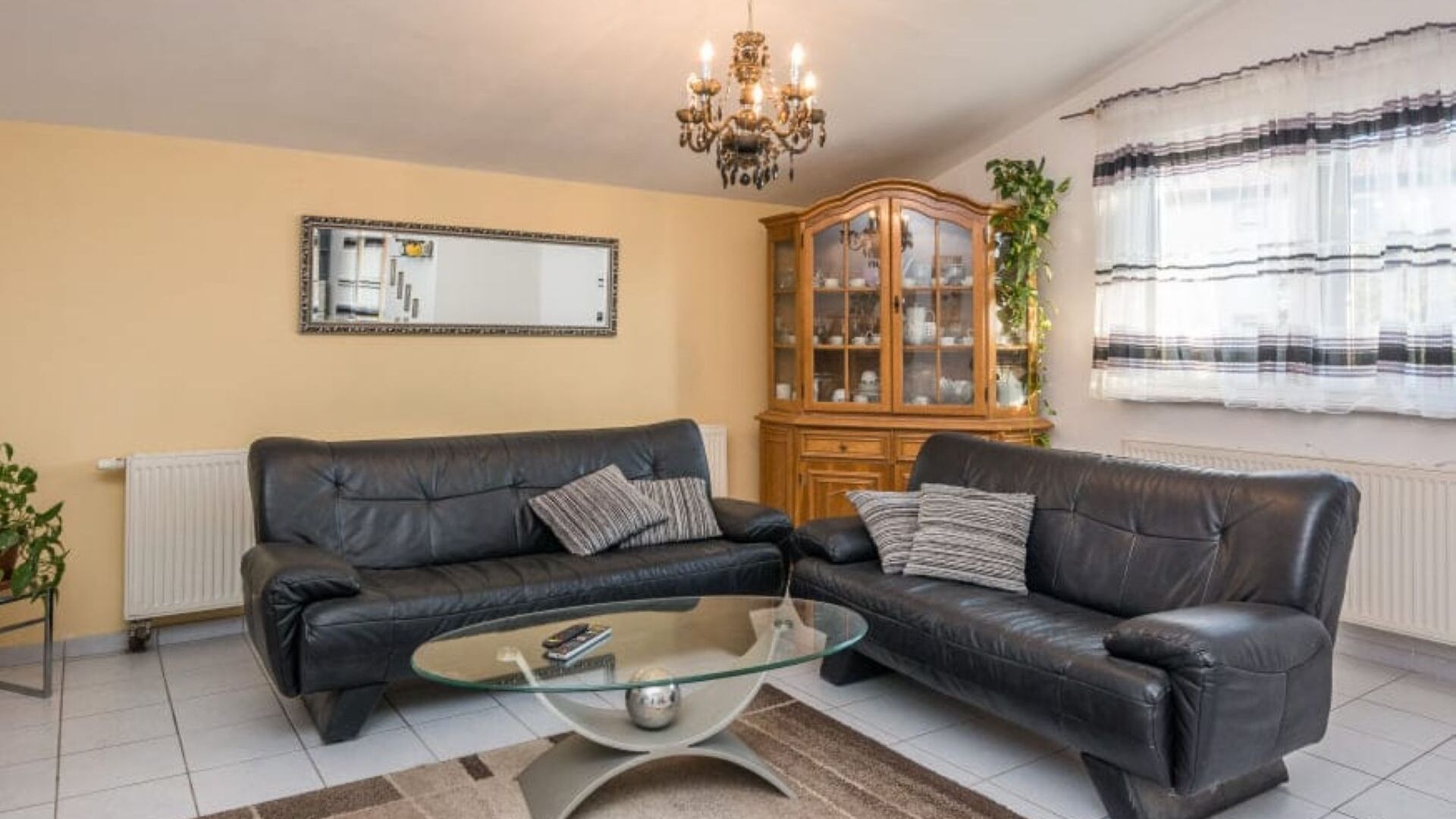Een woonkamer met twee zwarte leren banken, een glazen salontafel, een houten kast, een spiegel aan de muur en een radiator.