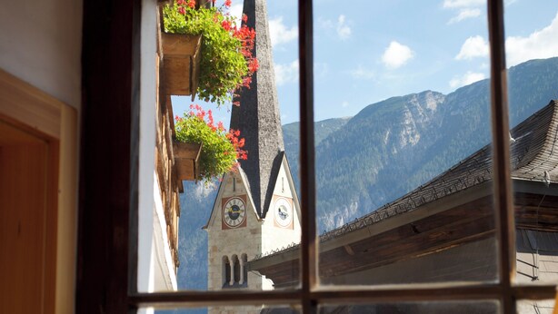 A window view of a building with a clock tower, a mountain backdrop, and a balcony with red flowers. - Pension Leopoldine (Hallstatt)