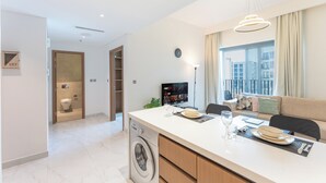 A modern kitchen with a washing machine, a dining area with a table set for two, and a living area with a sofa and a TV. - PVH - Azizi Mirage 1 - Tranquil 1 BR (Dubai)