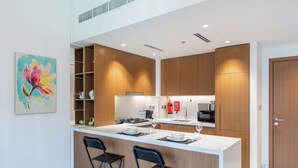 A modern kitchen with wooden cabinets, a white countertop, and two bar stools. - PVH - Azizi Mirage 1 - Tranquil 1 BR (Dubai)