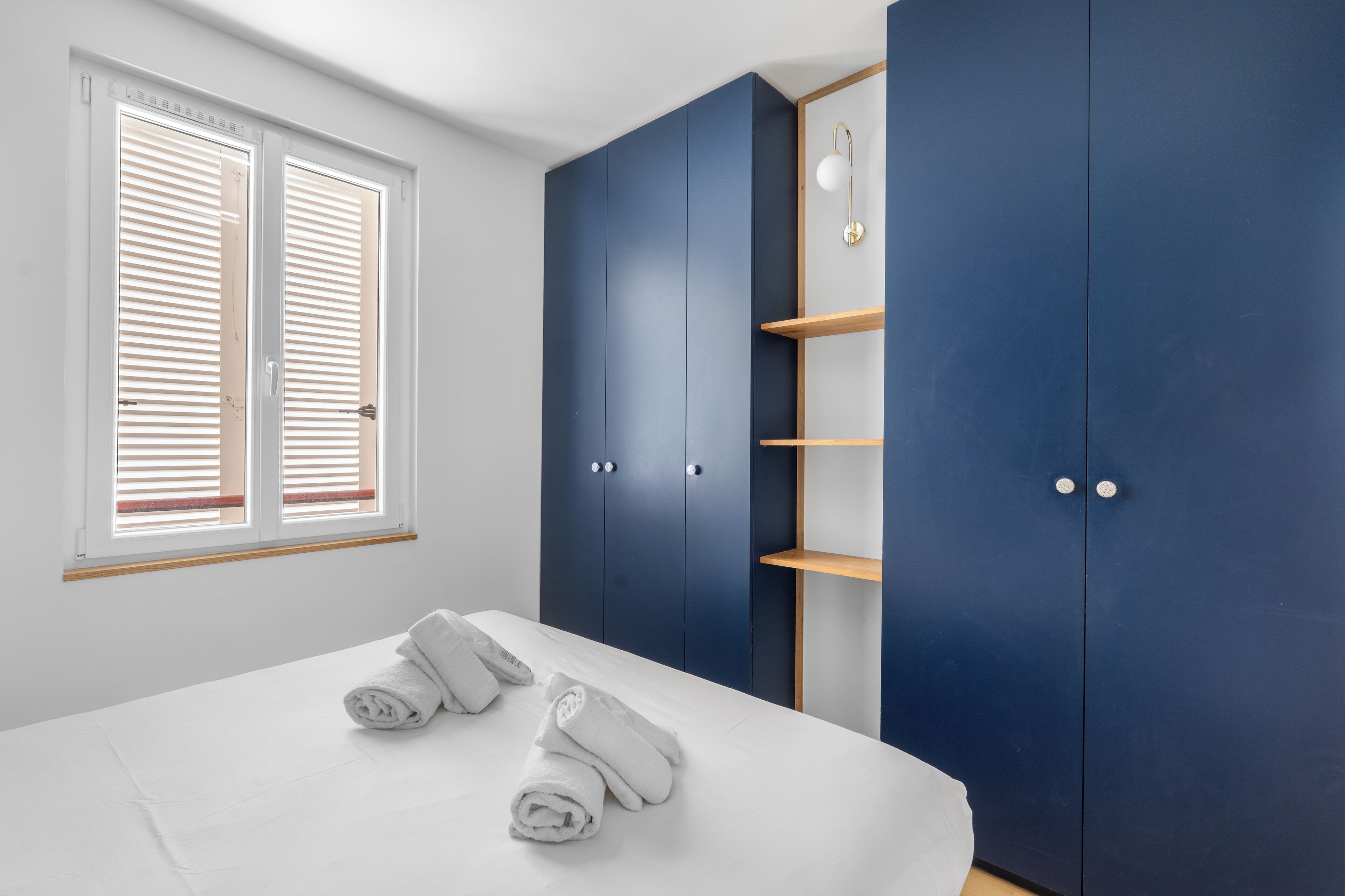 Une chambre à coucher avec un lit, deux serviettes enroulées, une armoire et une fenêtre avec des stores.