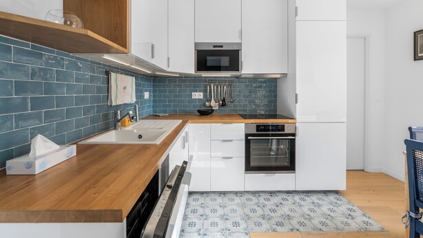 Een moderne keuken met witte kasten, een houten aanrechtblad, een blauwe betegelde spatwand en een vloerkleed met een patroon.