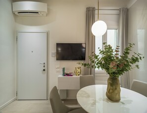 Quarto de hotel moderno com porta branca, TV de tela plana montada, mesa de console branca com um vaso de flores e uma mesa de jantar redonda com cadeiras.