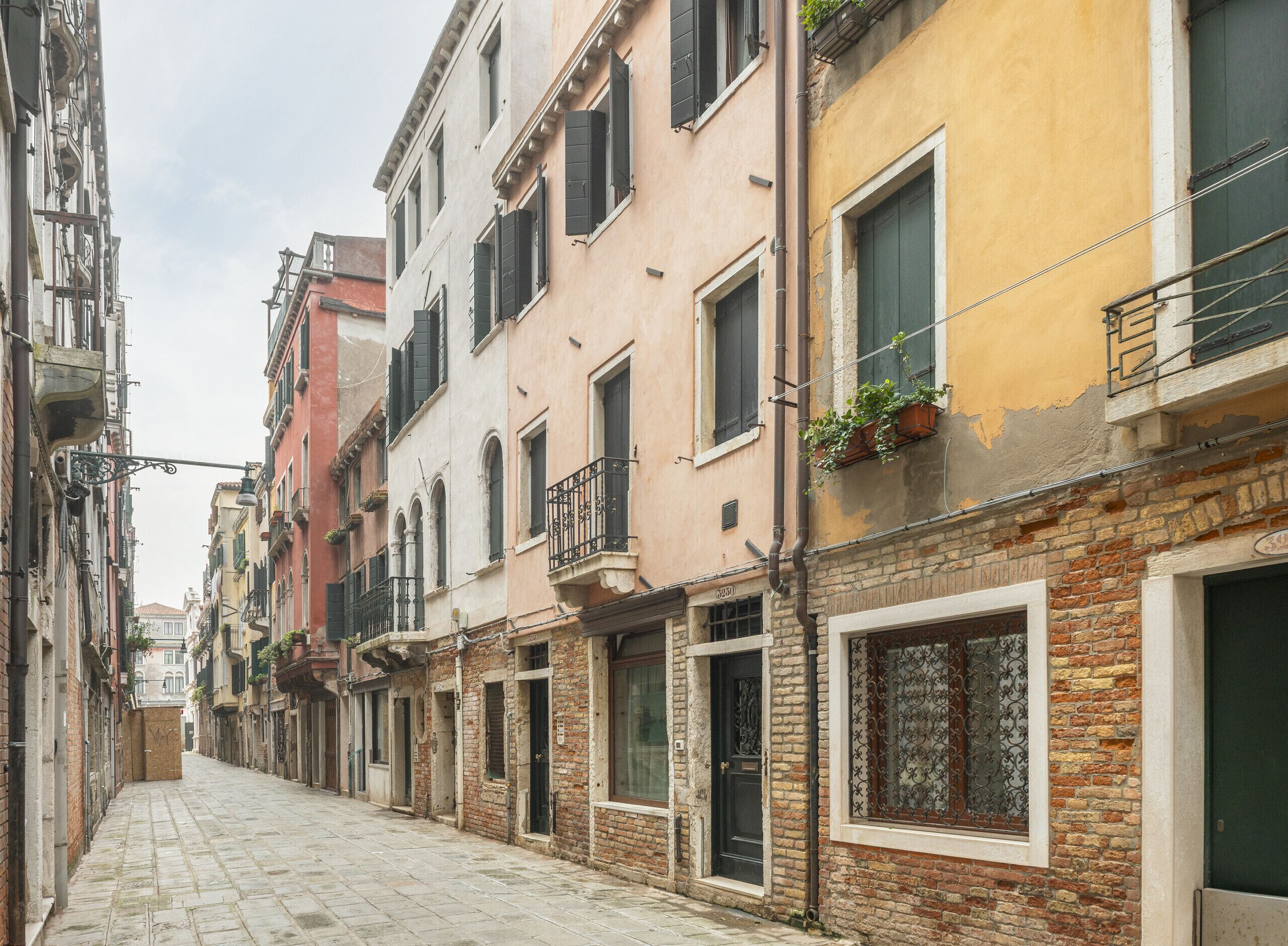 A narrow street lined with historic buildings, featuring balconies with plants, a cobblestone path, and a brick facade.