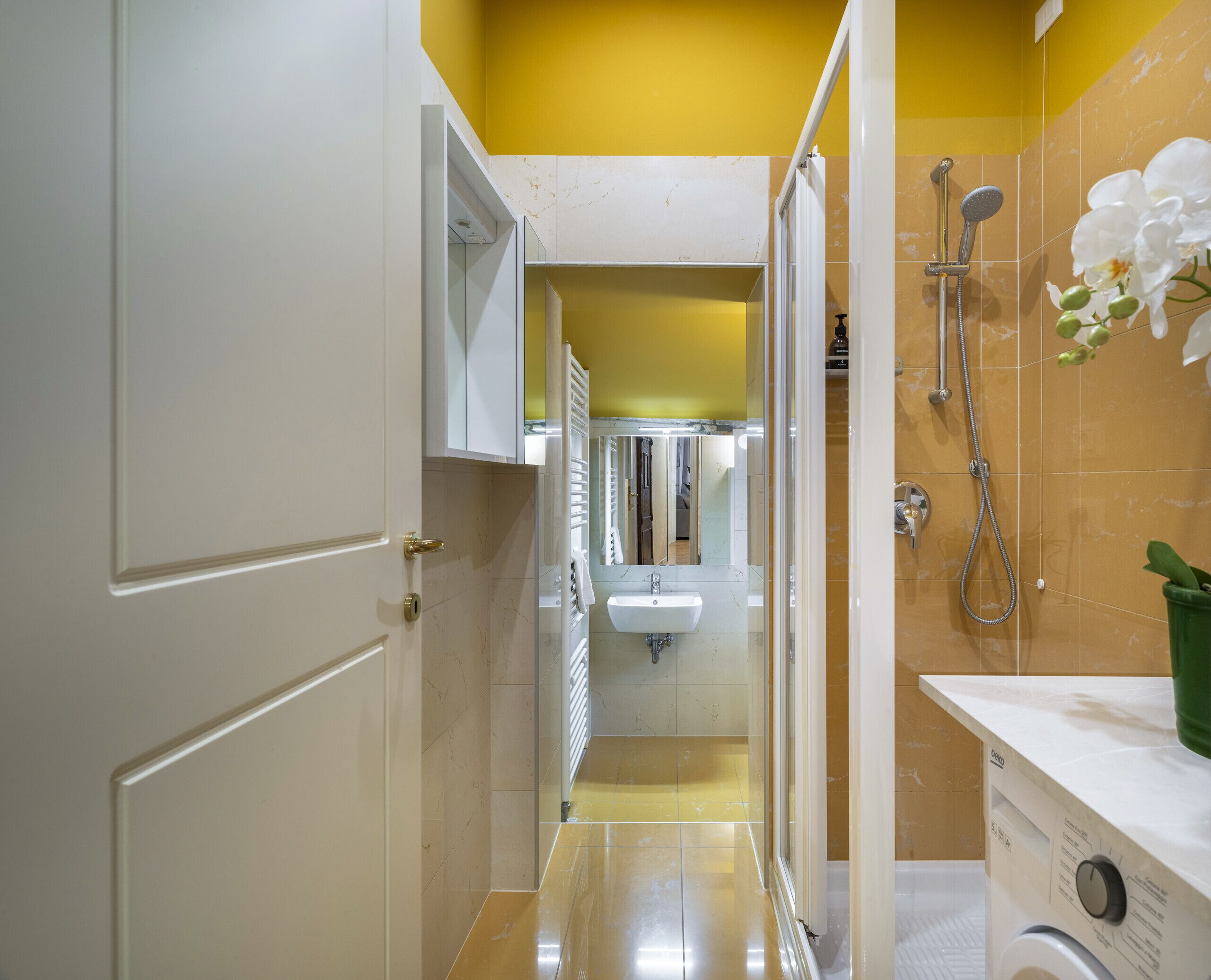 A narrow bathroom with a yellow wall, a white door, a glass shower enclosure, a sink, and a washing machine.