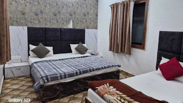 A bedroom with a large bed, a smaller bed, a nightstand, an air conditioning unit, and a window with curtains. - Shri Krishna Niwas (Mathura)