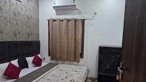 A bedroom with a bed, a headboard, a window with curtains, an air conditioning unit, and a door. - Shri Krishna Niwas (Mathura)
