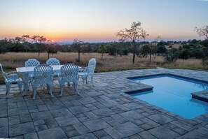 Een zwembad met uitzicht op de ondergaande zon, omringd door een terras met stoelen en een tafel.
