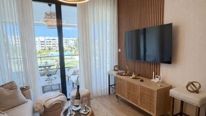 Ett modernt vardagsrum med en stor TV, en soffa, ett soffbord och utsikt över en pool och byggnader.