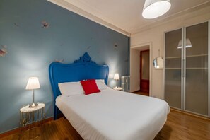 A bedroom with a blue headboard, white bedding, a red pillow, two bedside lamps, and a wardrobe with a mirror. - GuestHost - Cozy 90m2 apartment, perfect for 4 people, located on the ground floor of a typical building with cloister.The accommodation is located in the Re di Roma/San Giovanni district, a lively and central residential and commercial area.The city's (Rome)