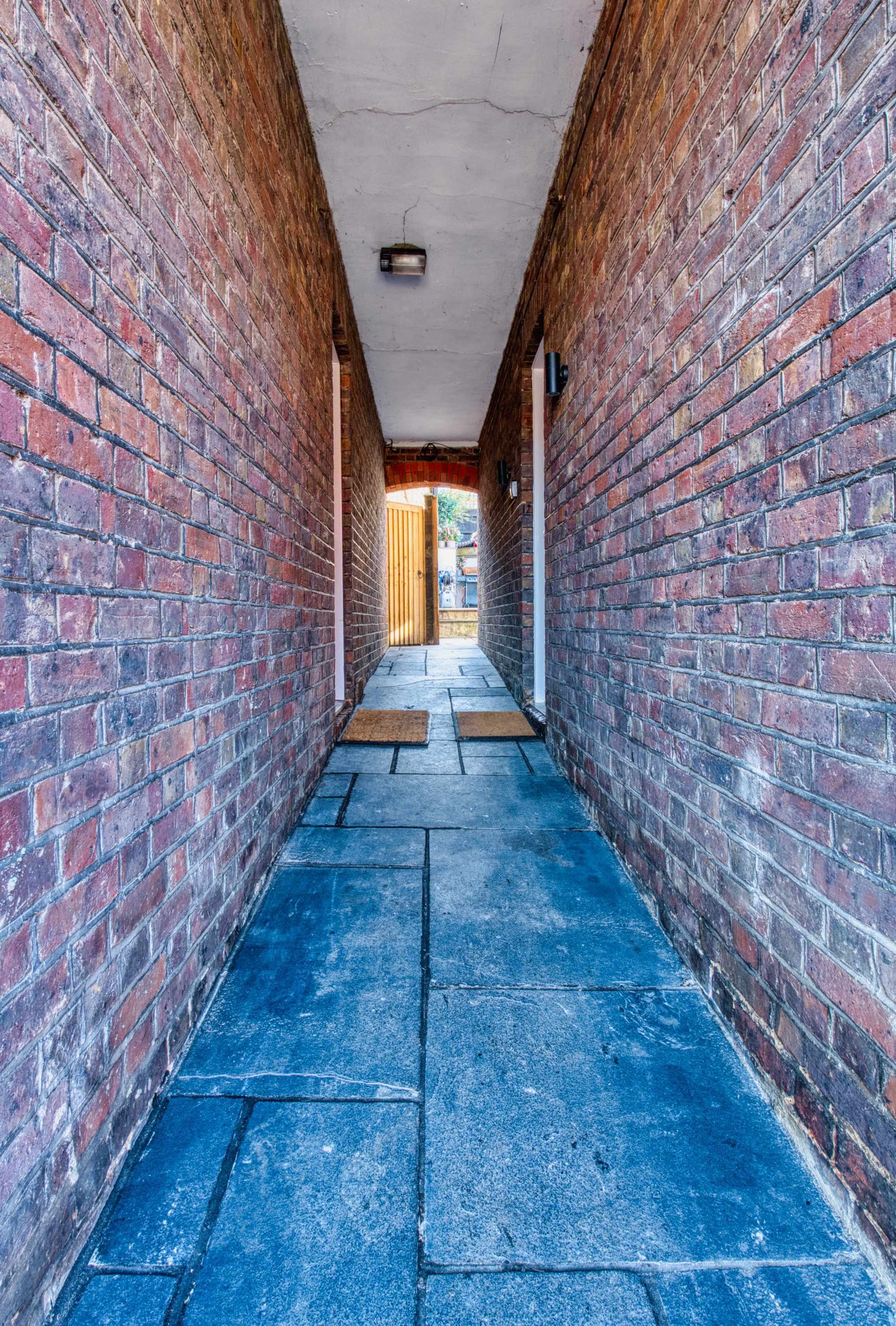 A narrow brick corridor with a blue tiled floor and a wooden door at the end.