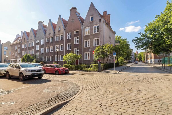 Eine Kopfsteinpflasterstraße gesäumt von traditionellen Gebäuden, parkenden Autos und wolkenlosem, blauem Himmel.