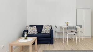 Un soggiorno moderno con un divano blu scuro, cuscini decorati, un piccolo tavolino con un vaso, una scrivania bianca con una sedia e una lampada a sospensione grigia.