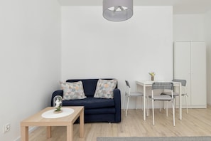 Ein modernes Wohnzimmer mit einem dunkelblauen Sofa, gemusterten Kissen, einem kleinen Holztisch mit einer Vase, einem weißen Schreibtisch mit Stuhl und einer grauen Pendelleuchte.