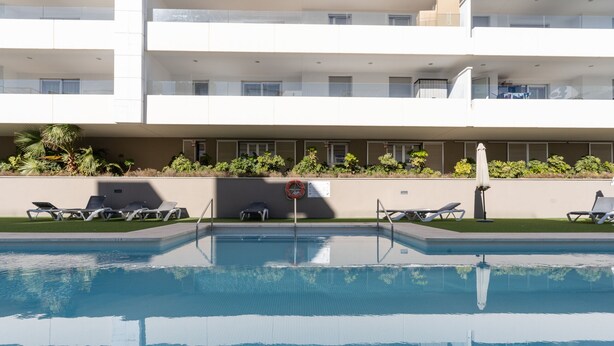 A swimming pool with lounge chairs, a lifeguard stand, and a building with balconies and windows. - Acqua by Time & Home (San Pedro de Alcántara)