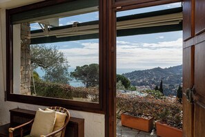 A room with a large window offering a view of a hillside and a body of water. There is a wicker chair and a wooden table in the room. - La Briciola, sea view, garden, private parking (Santa Margherita Ligure)