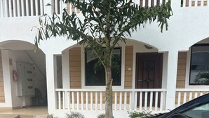 Ein Gebäude mit weißer Fassade, einem Balkon mit weißen Geländern und einem gepflasterten Eingangsbereich mit Baum und geparktem Auto.