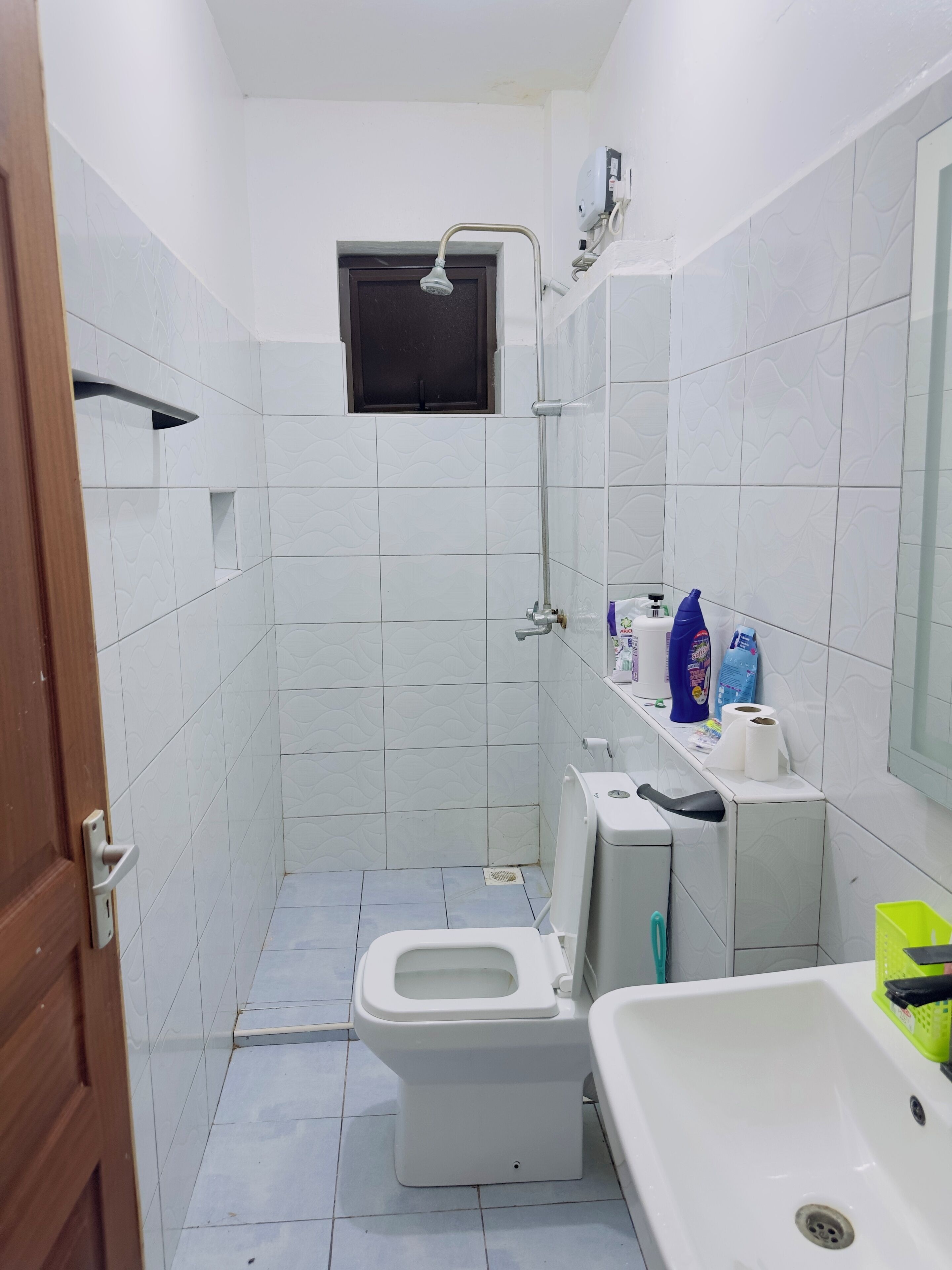 A small, compact bathroom with white tiled walls, a toilet, a sink, and a shower. There is a window above the toilet and a shelf with toiletries.