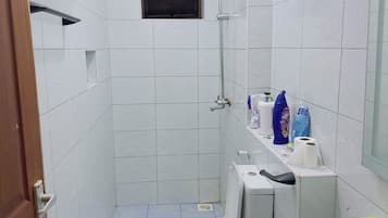 A small, compact bathroom with white tiled walls, a toilet, a sink, and a shower. There is a window above the toilet and a shelf with toiletries.